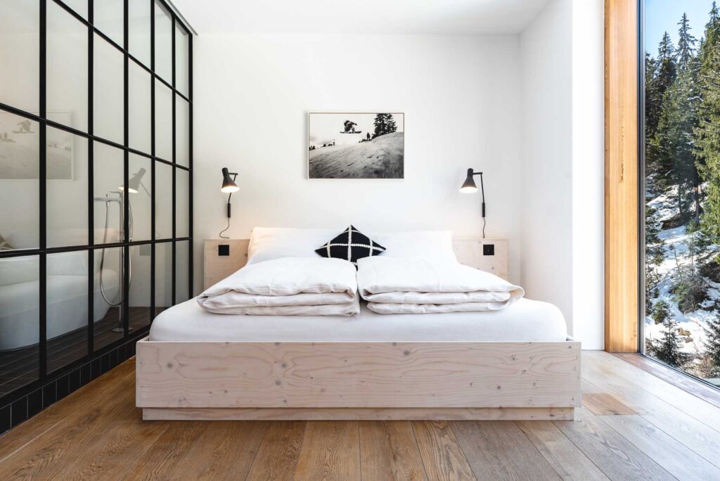 Modernes Designzimmer im Riders Hotel Laax Helles Doppelzimmer im Riders Hotel Laax mit modernem Interior, grossem Fenster und Blick in den verschneiten Wald – fotografiert vom Hotelfotograf Christian Meixner.